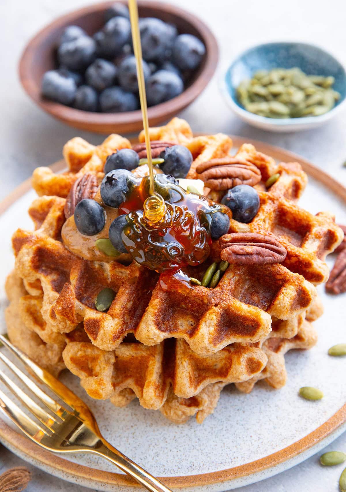 Three waffles stacked on a plate topped with almond butter, blueberries, and pecans with honey being poured on top. A small bowl of blueberries and a small bowl of pumpkin seeds in the background. There's a golden fork next to the waffles and a golden napkin to the side.