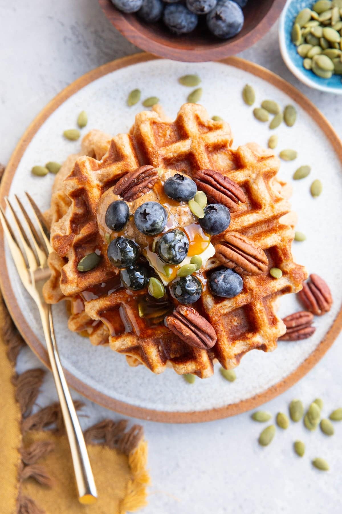 Plate of waffles with almond butter, regular butter, blueberries, pumpkin seeds and pecans on top. A gold fork to the side and bowls of blueberries and pumpkin seeds.