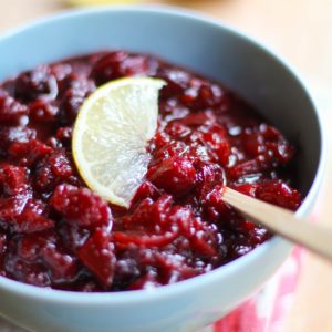Cranberry Sauce with Ginger and Maple recipe inside of a blue bowl sitting on top of a pretty napkin and a cutting board.
