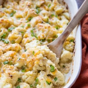 Casserole dish full of cheesy cauliflower casserole with a red napkin to the side and a wooden spoon ready to serve.
