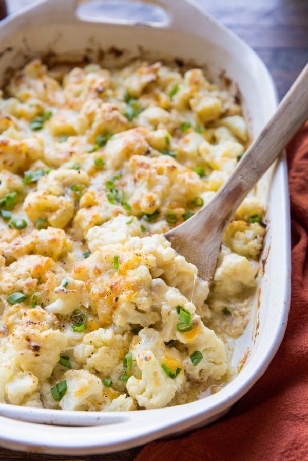 Casserole dish full of cheesy cauliflower casserole with a red napkin to the side and a wooden spoon ready to serve.