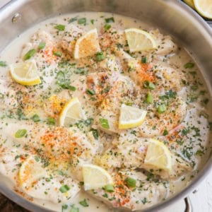 Creamy lemon garlic chicken in a stainless steel skillet with chopped green onions and parsley on top. A bowl of fresh lemons in the background and a bowl of chopped green onions, and a small bowl of dried parsley.