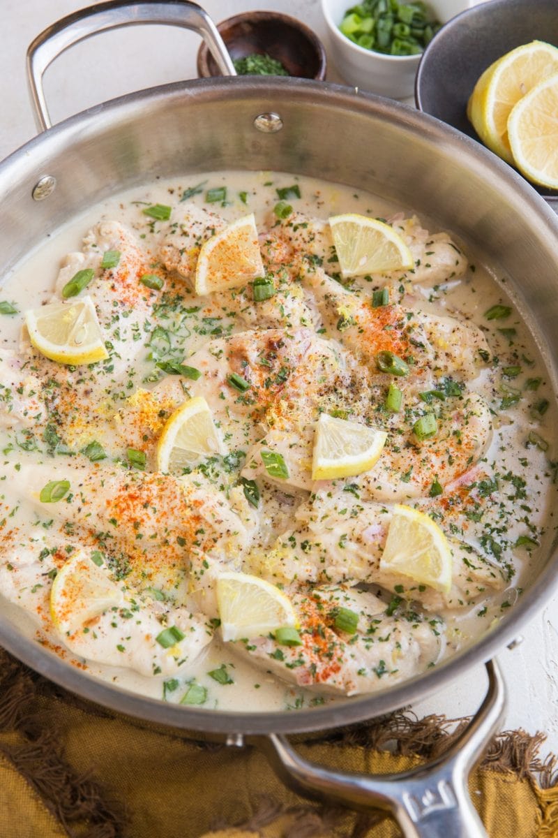 Creamy lemon garlic chicken in a stainless steel skillet with chopped green onions and parsley on top. A bowl of fresh lemons in the background and a bowl of chopped green onions, and a small bowl of dried parsley.