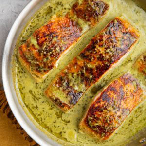 Stainless steel skillet with seared salmon filets and a creamy pesto sauce.