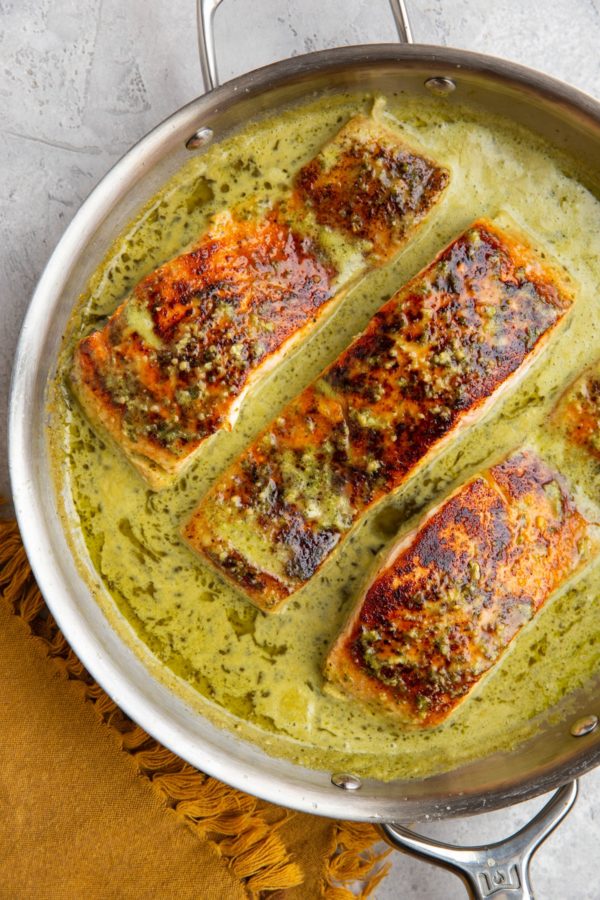 Stainless steel skillet with seared salmon filets and a creamy pesto sauce.