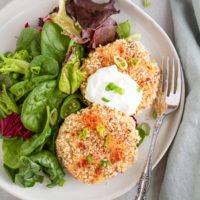 Plate with two cauliflower potato cakes topped with yogurt dipping sauce and a side salad.