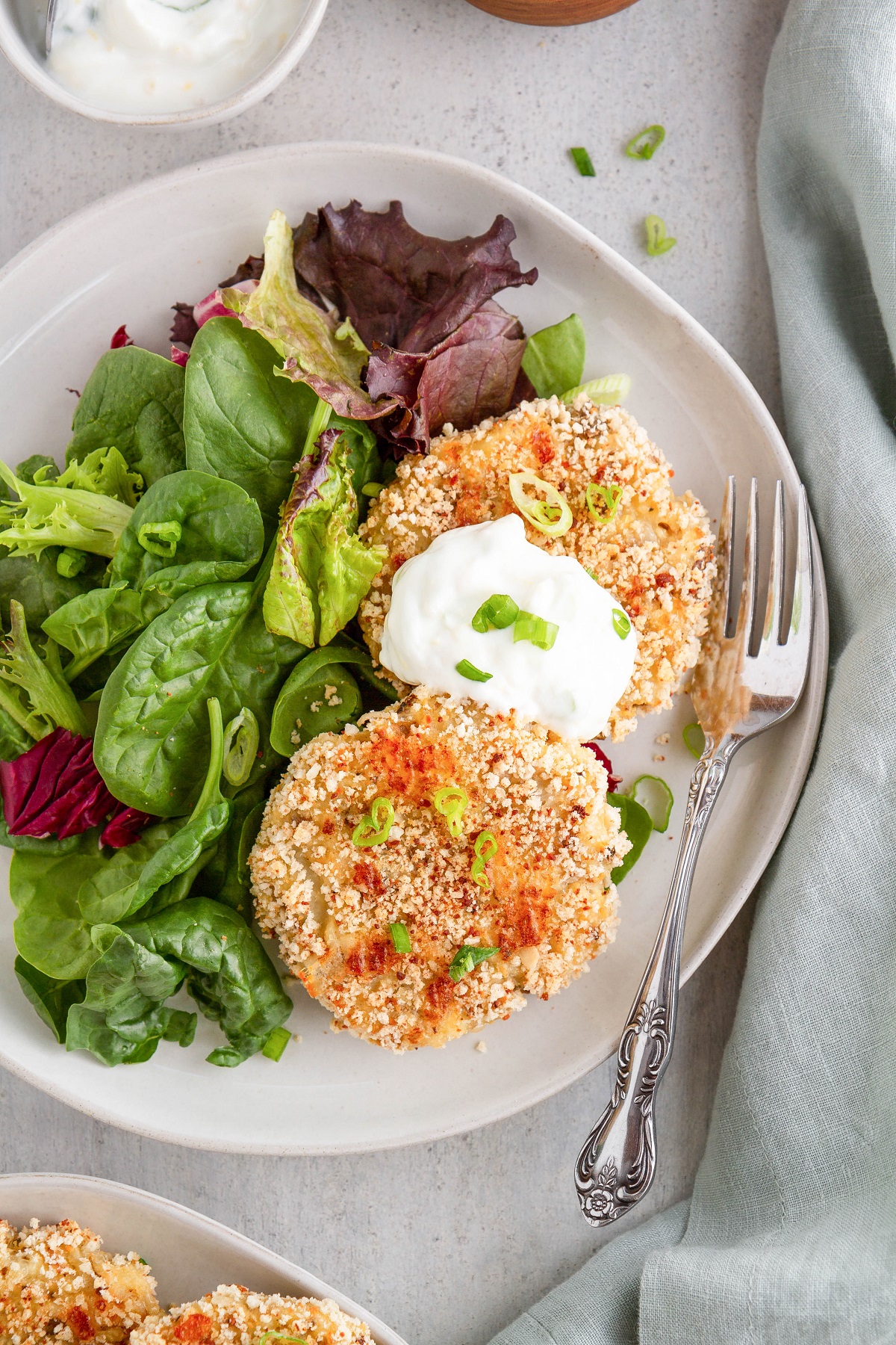 Plate with two cauliflower potato cakes topped with yogurt dipping sauce and a side salad.