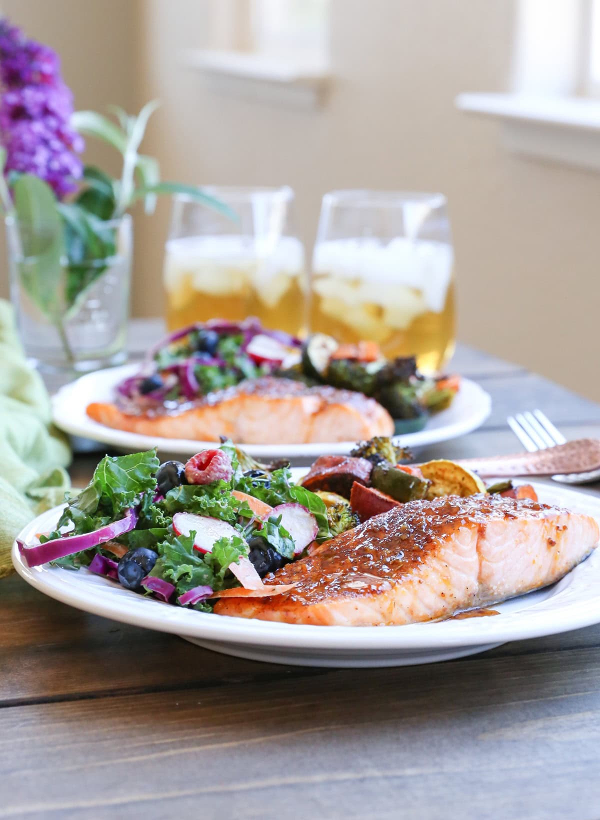 Two plates of Crispy Honey-Glazed Salmon with roasted vegetables and kale salad with glasses of iced tea in the background. Ready to eat.