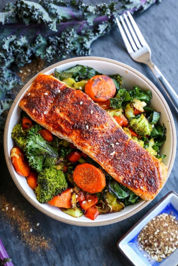Bowl of crispy salmon on top of roasted vegetables with a fresh kale leaf to the side.