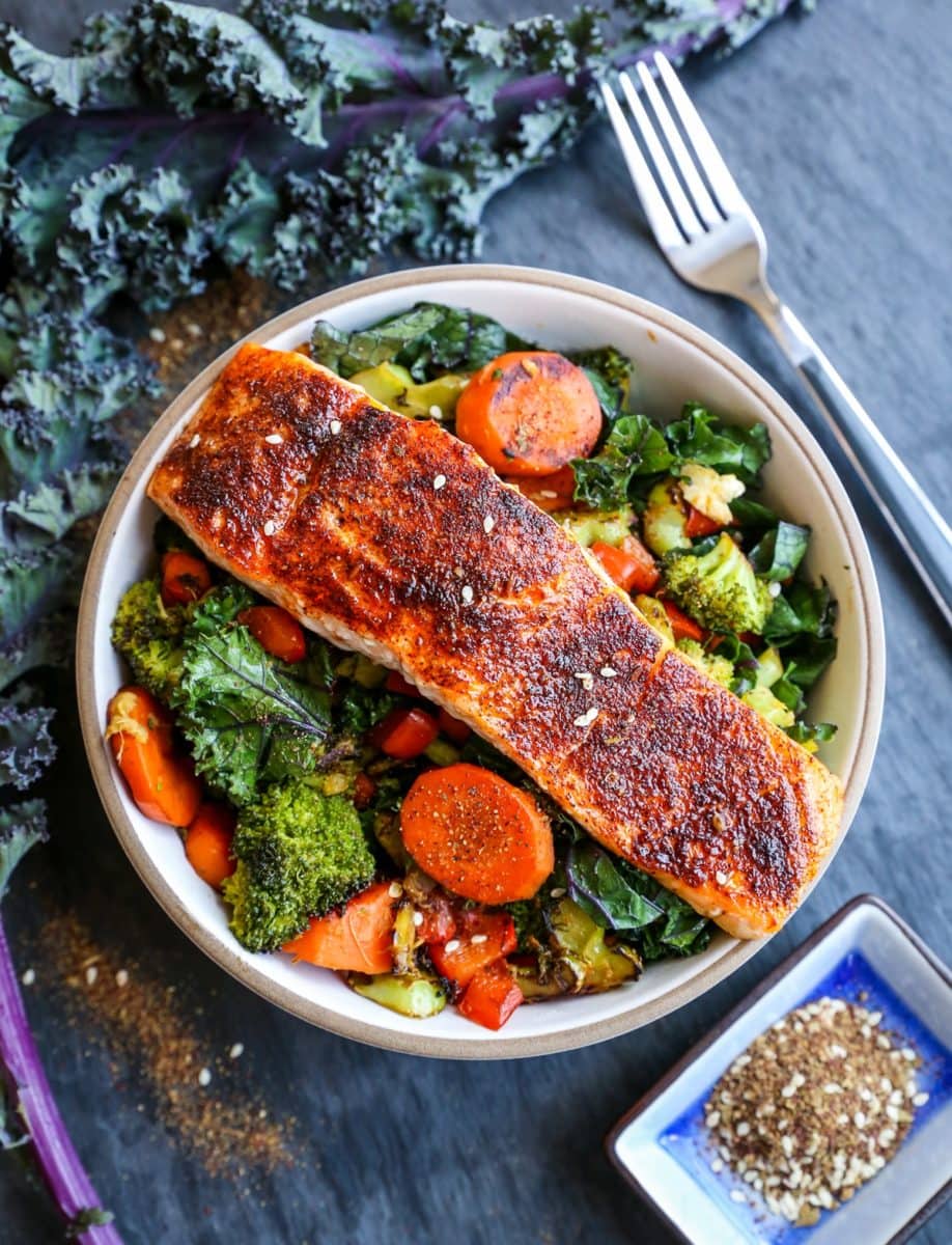 Bowl of crispy salmon on top of roasted vegetables with a fresh kale leaf to the side.