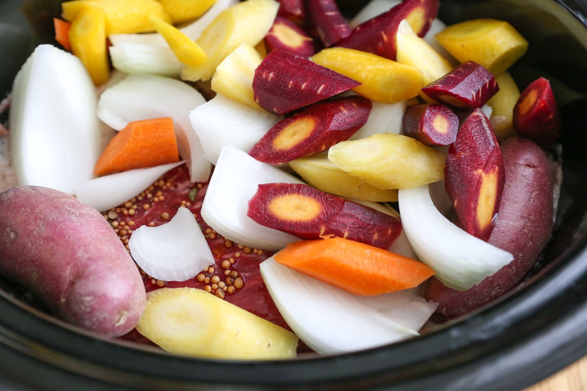 Slow cooker full of raw corned beef, onions, chopped carrots, and potatoes, ready to slow cook.