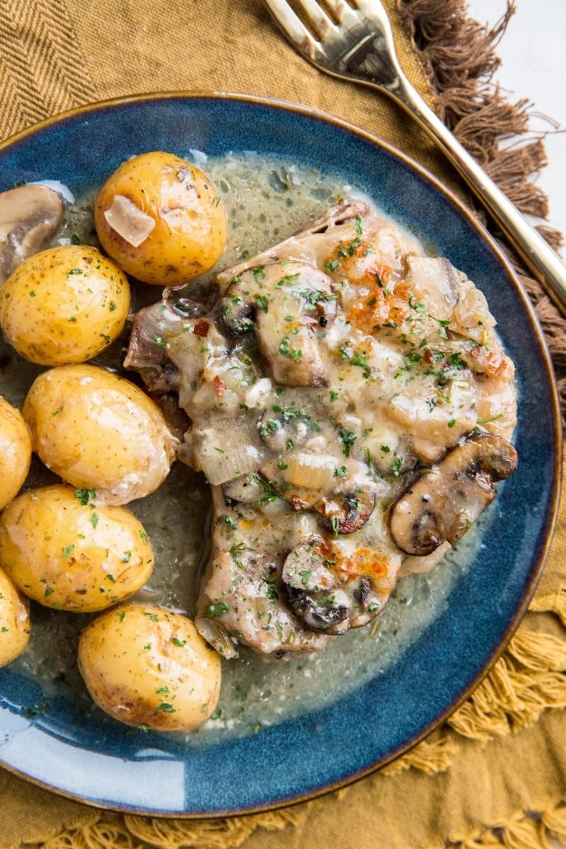 Blue plate with a Pork Chop with creamy garlic sauce, potatoes, and mushrooms with a golden napkin and a gold fork, ready to serve.