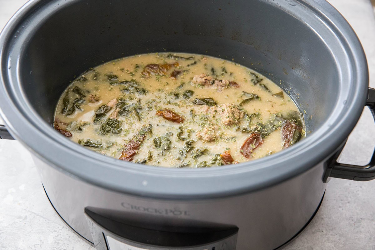 A slow cooker filled with a creamy sausage soup containing pieces of meat, leafy greens, and sun-dried tomatoes simmers gently on a light-colored countertop.