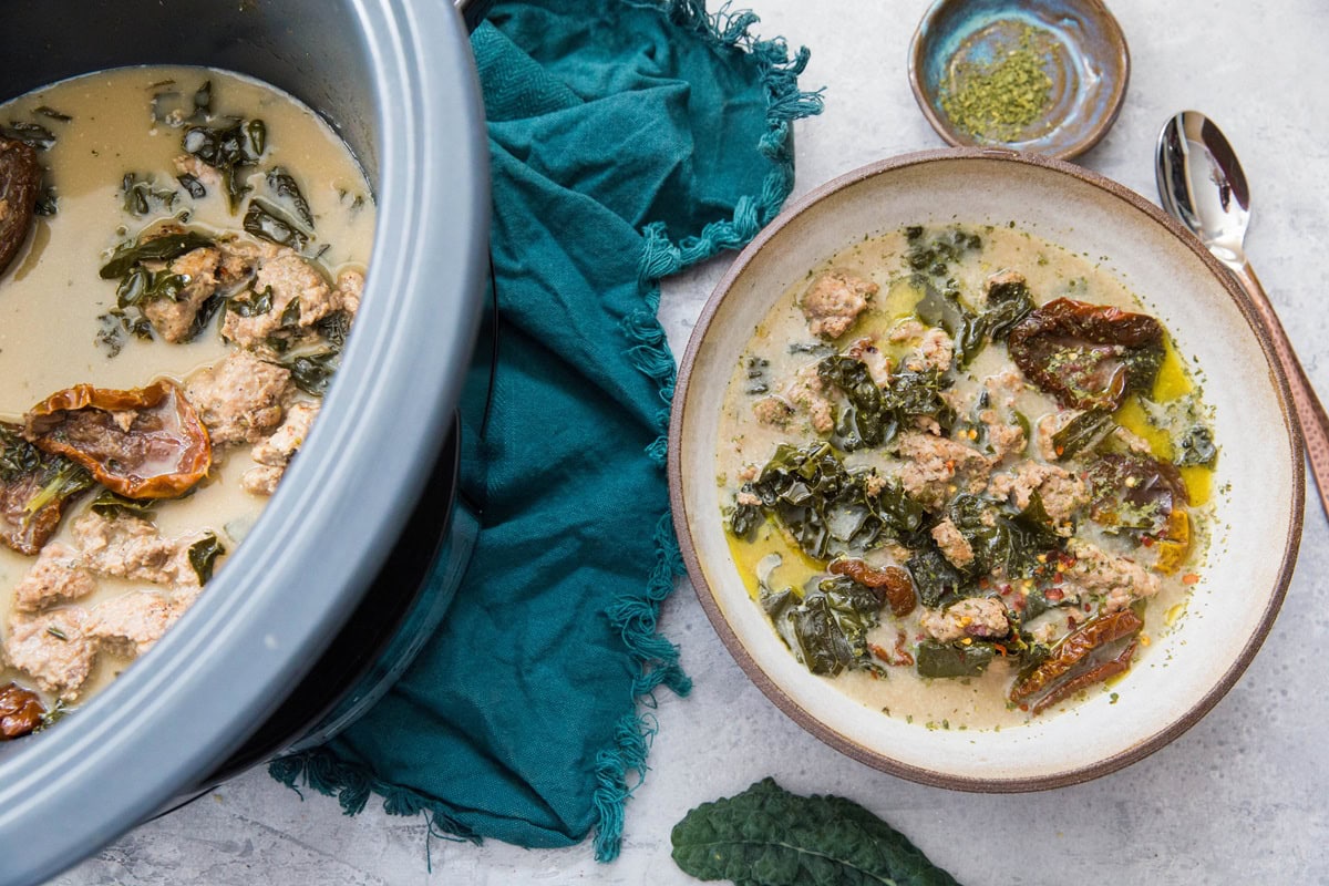 A bowl of creamy sausage soup with pieces of chicken, kale, and sun-dried tomatoes sits beside a slow cooker filled with more soup. A green cloth, a spoon, and a small dish of herbs rest on the light surface nearby.