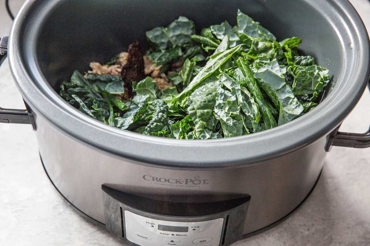A slow cooker filled with leafy kale and ground meat, perfect for a hearty sausage soup, sits on a light-colored countertop.