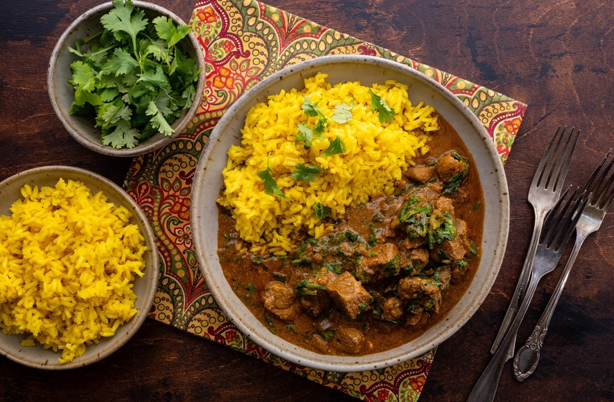 Bowl of lamb curry with saffron rice with a bowl of rice and a bowl of cilantro to the side.