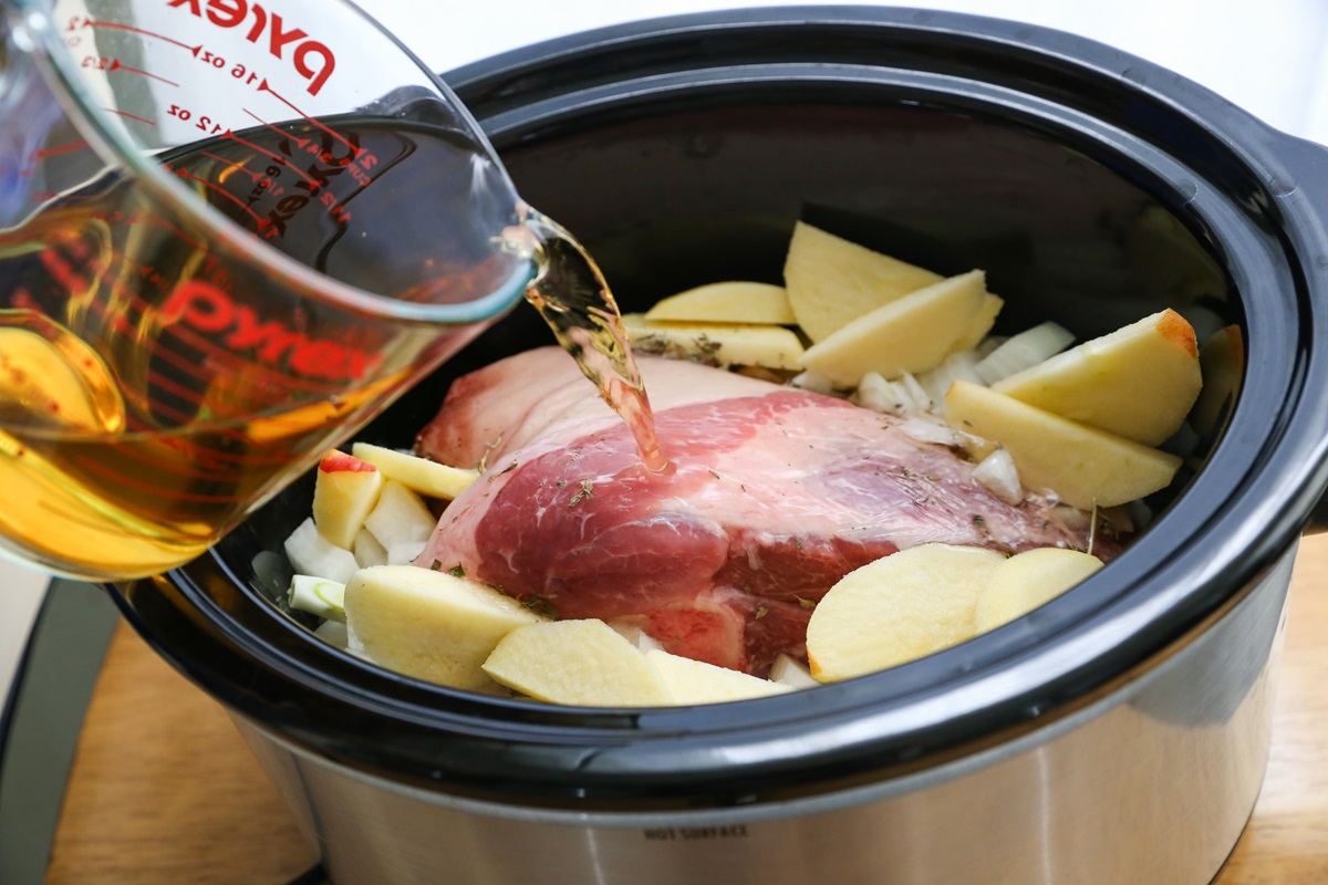 Pouring liquid into a slow cooker with pork butt, apples, and onions.