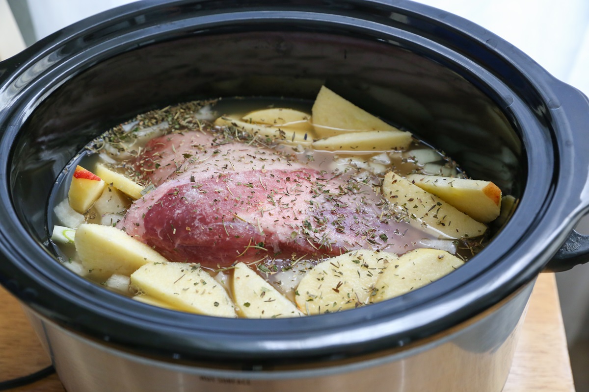 Slow cooker with pork shoulder roast, apples, onions, seasonings, and broth, ready to be slow cooked.