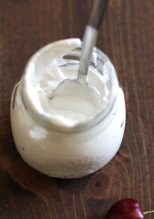 A glass jar filled with creamy white yogurt sits on a wooden surface, topped with a swirl of coconut whipped cream, with a metal spoon inside. A single red cherry lies nearby on the table.