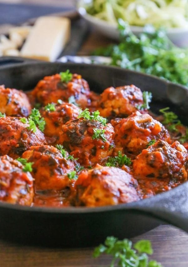 Cast iron skillet full of turkey meatballs slathered in tomato sauce. Fresh parsley to the side and parmesan cheese in the background.