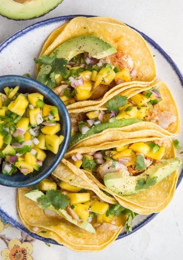 Three Fish Tacos with Mango Salsa and fresh cilantro on corn tortillas sitting on a blue-rimmed plate with a floral napkin to the side and a bowl of fresh mango salsa.