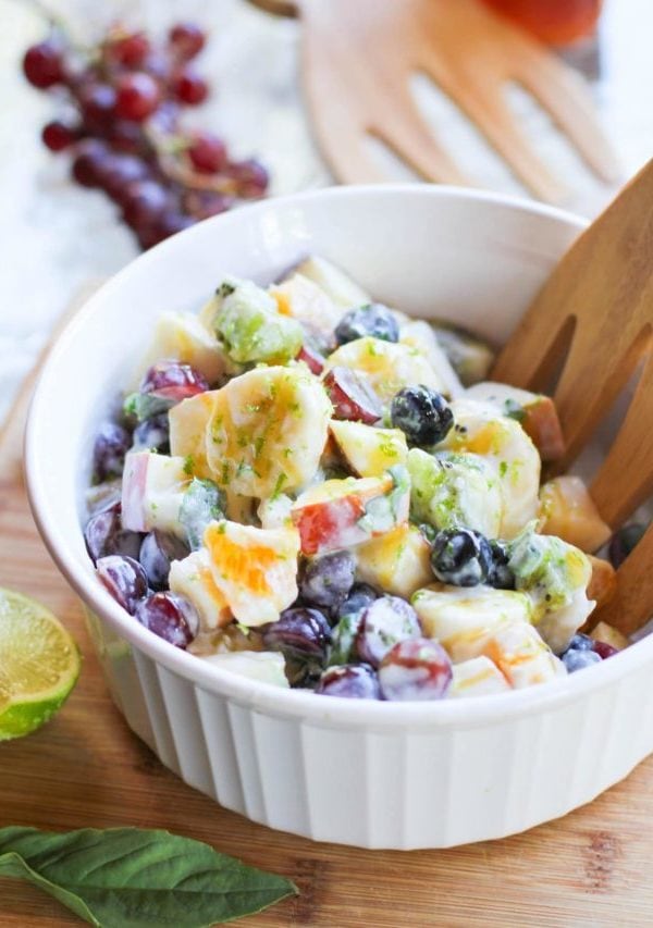White bowl of fruit salad with a bunch of grapes in the background and a fresh basil leaf in front of the bowl.