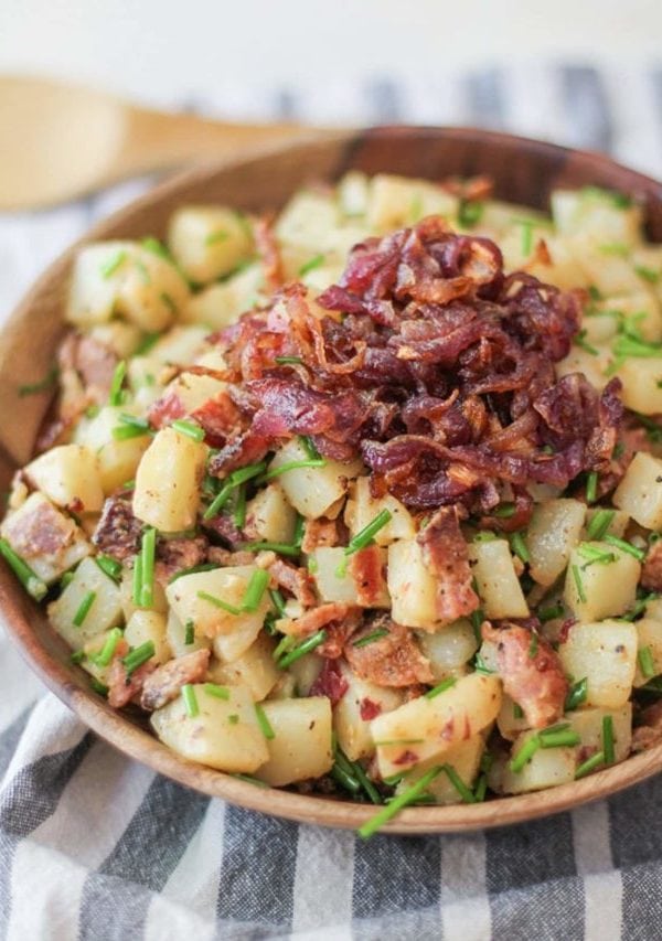 German-Style Potato Salad with bacon, caramelized onions, and chives in a bowl with the caramelized onions on top. The wooden bowl is on top of a striped napkin and the dish is ready to serve.