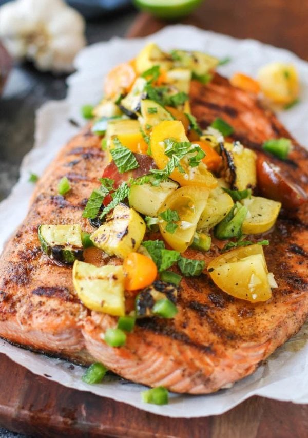 large piece of grilled salmon on a piece of parchment paper on a cutting board with grilled zucchini, tomatoes, green onions and fresh herbs on top. Ready to serve.