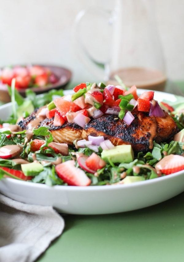 White bowl with grilled salmon on top of salad with strawberry salsa and homemade balsamic vinaigrette with a napkin to the side and balsamic vinaigrette in the background.