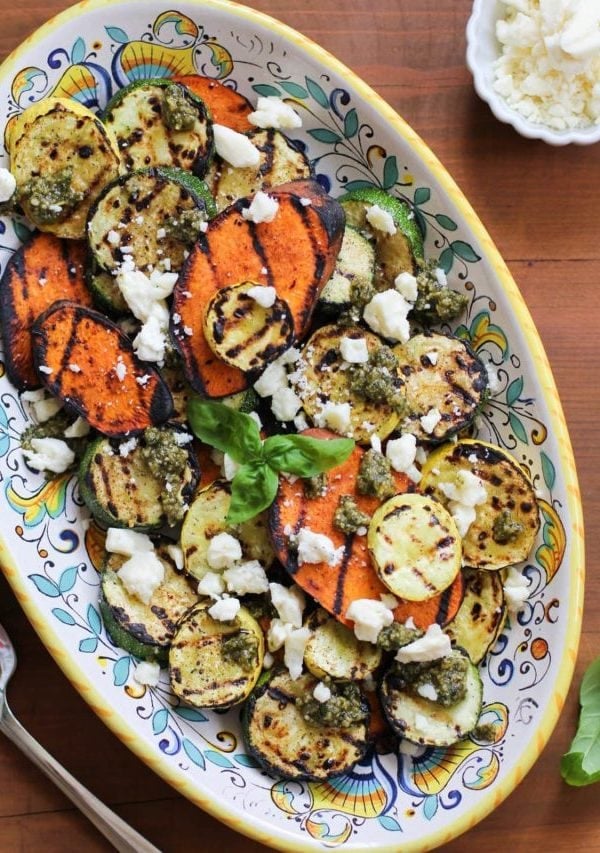 Platter of grilled sweet potatoes, yellow squash, and zucchini with feta cheese sprinkled on top and pesto sauce drizzled over everything.