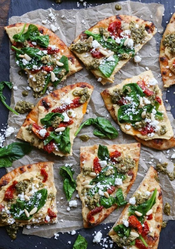 Black slate with slices of hummus flatbread on top, topped with sun-dried tomatoes, pesto sauce, spinach, and more.