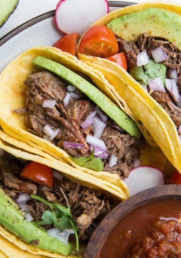 Three shredded beef tacos on a plate with tomato salsa, avocado, cilantro, and red onion. Ready to eat.