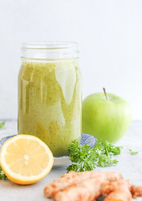 Large glass of smoothie with a green apple in the background, fresh parsley, a lemon and turmeric.