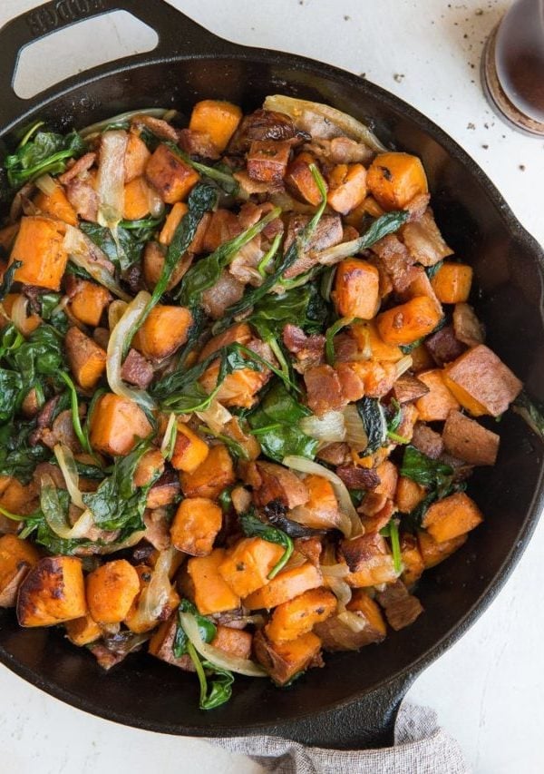 Sweet Potato Hash Recipe in a skillet with crispy bacon, onions, and spinach and a linen wrapped around the handle of the skillet. Pepper grinder to the side.