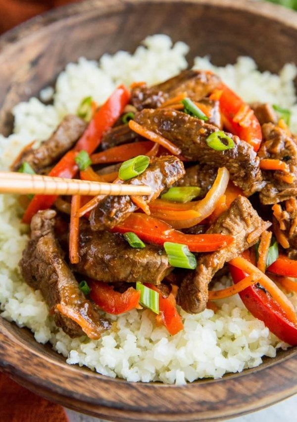 Szechuan beef in a large wooden bowl on top of cauliflower rice with a pair of chopsticks taking a bite of beef.