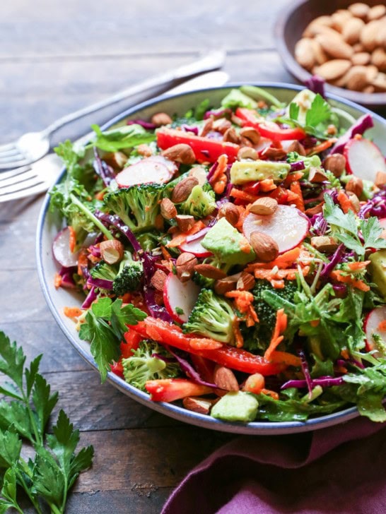 Big bowl of salad with fresh vegetables, almonds, avocado and lemon-parsley dressing. A purple napkin and fresh parsley to the side.