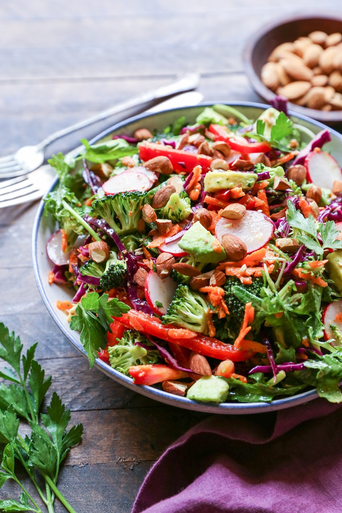 Big bowl of salad with fresh vegetables, almonds, avocado and lemon-parsley dressing. A purple napkin and fresh parsley to the side.