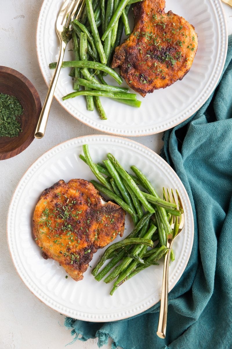 Two plates with cooked air fryer pork chops served with cooked green beans. Golden forks for eating and a blue napkin to the side.