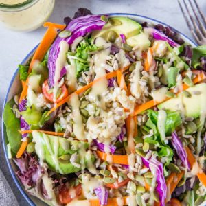 large bowl of summer salad recipe with greens, carrots, cabbage, avocado, brown rice and homemade dressing. Jar of dressing and bowl of pumpkin seeds to the side.