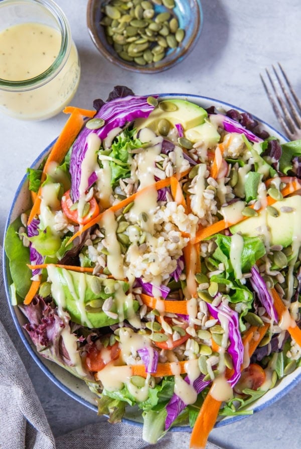large bowl of summer salad recipe with greens, carrots, cabbage, avocado, brown rice and homemade dressing. Jar of dressing and bowl of pumpkin seeds to the side.