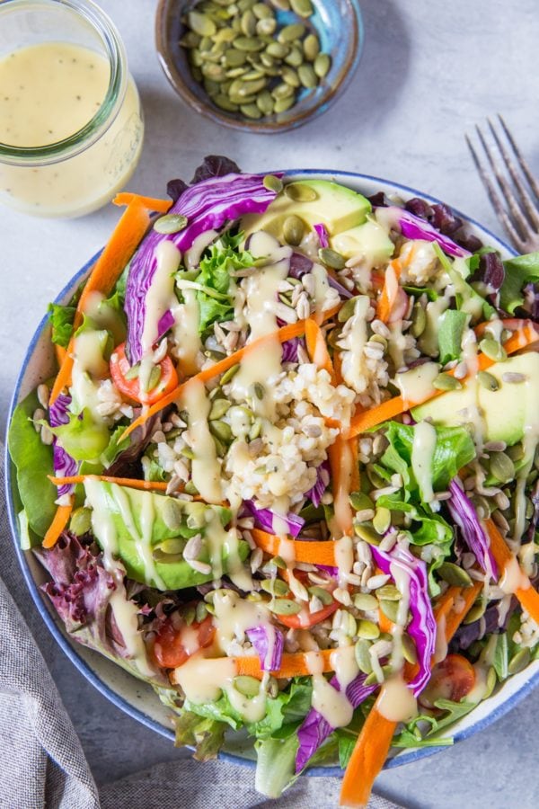 large bowl of summer salad recipe with greens, carrots, cabbage, avocado, brown rice and homemade dressing. Jar of dressing and bowl of pumpkin seeds to the side.