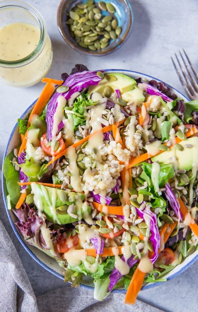 large bowl of summer salad recipe with greens, carrots, cabbage, avocado, brown rice and homemade dressing. Jar of dressing and bowl of pumpkin seeds to the side.