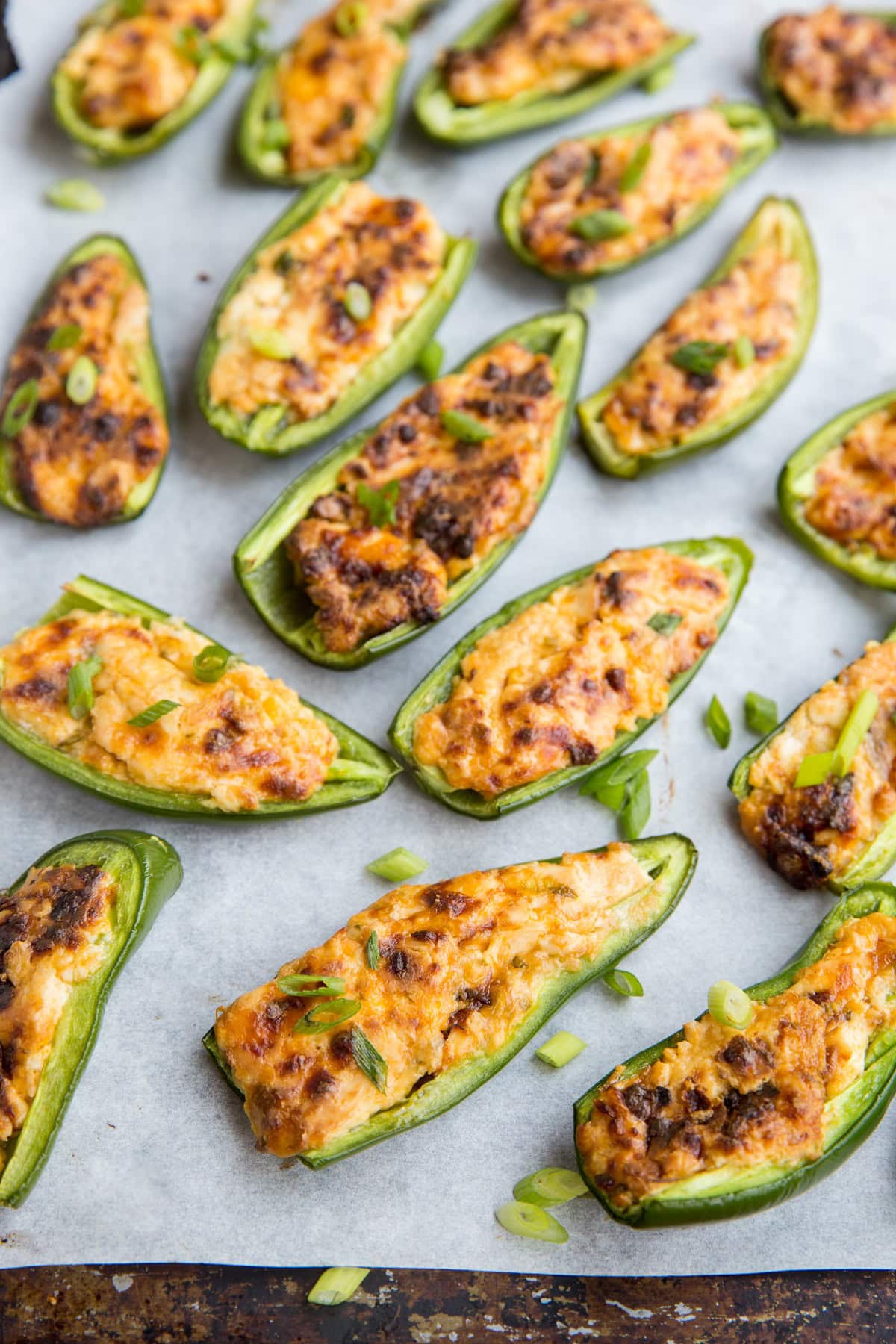 Jalapeños that have been sliced in half and stuffed with a creamy cream cheese mixture sitting on a sheet of parchment paper on a baking sheet and sprinkled with green onions. Golden brown and fresh out of the oven.