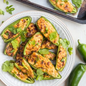 Plate full of cream cheese stuffed baked jalapeno halves with cilantro sprinkled on top. A baking sheet with more poppers and fresh jalapeno to the side with a bright floral napkin.
