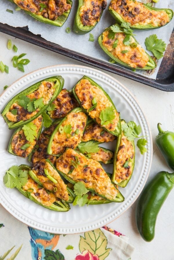 Plate full of cream cheese stuffed baked jalapeno halves with cilantro sprinkled on top. A baking sheet with more poppers and fresh jalapeno to the side with a bright floral napkin.
