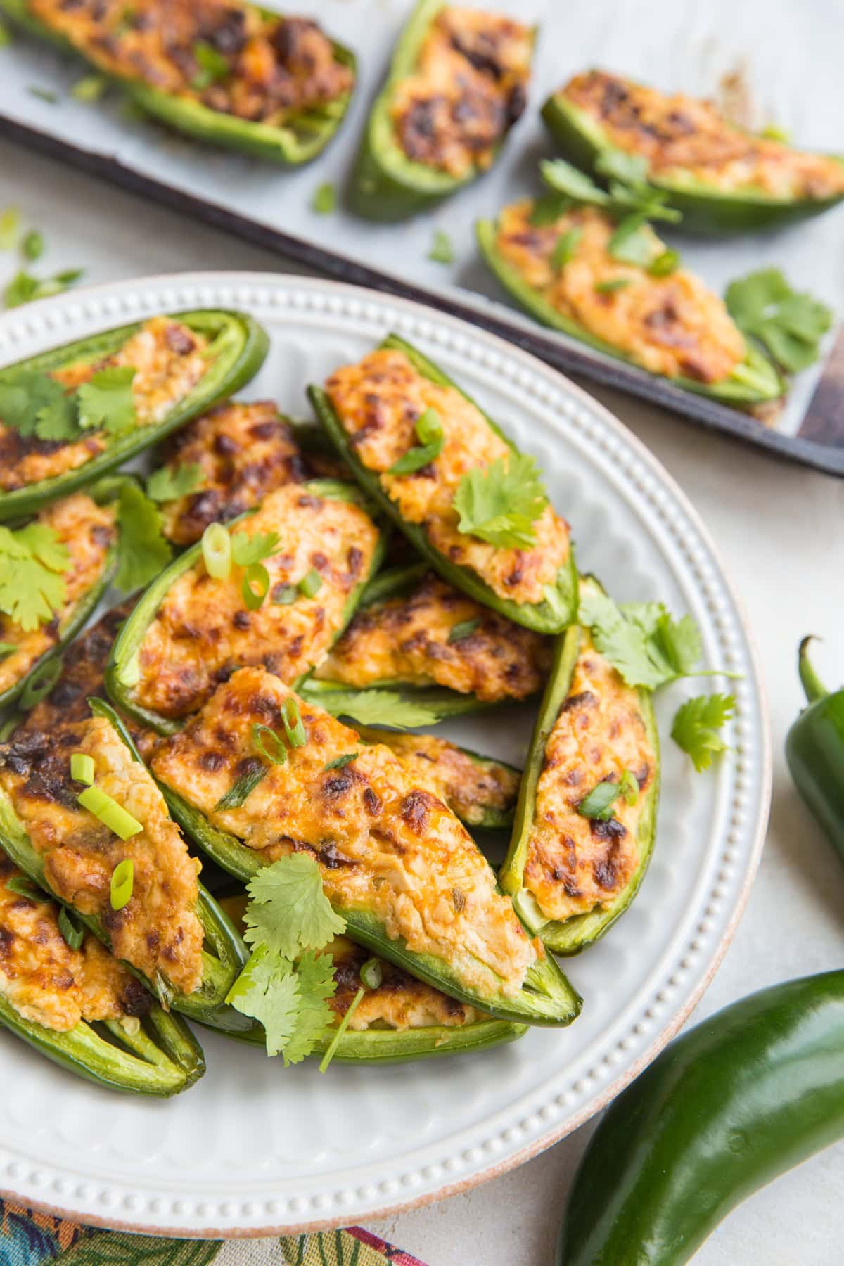 Jalapeno poppers on a white plate with the baking sheet of the rest of the poppers in the background, and a napkin and a fresh jalapeno to the side.