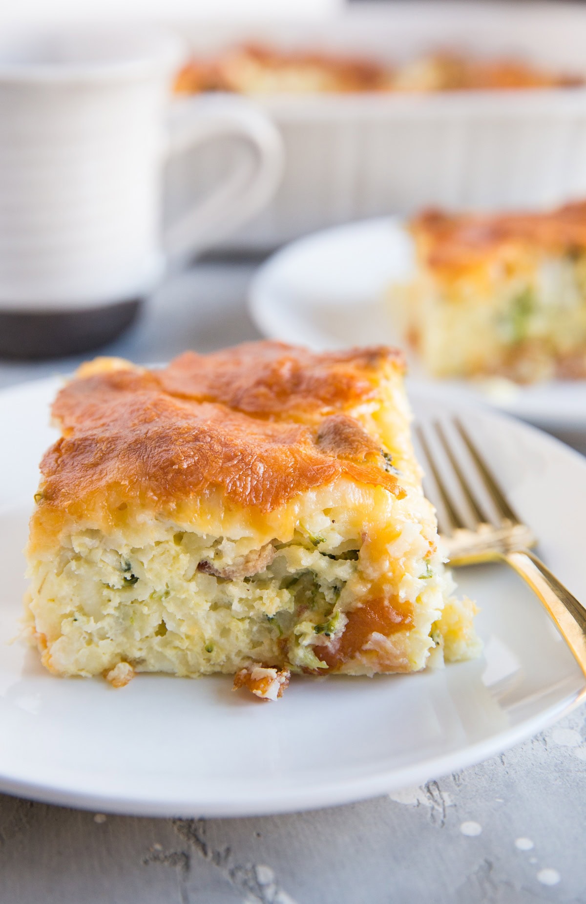 Slice of Broccoli Cheddar Egg Hashbrown Casserole on a white plate with a fork to the side. Another plate of hashbrown casserole in the background.
