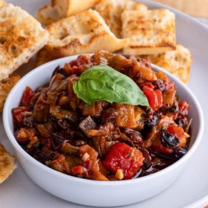 Bowl of eggplant caponata on a plate with toasted baguette, ready to serve.