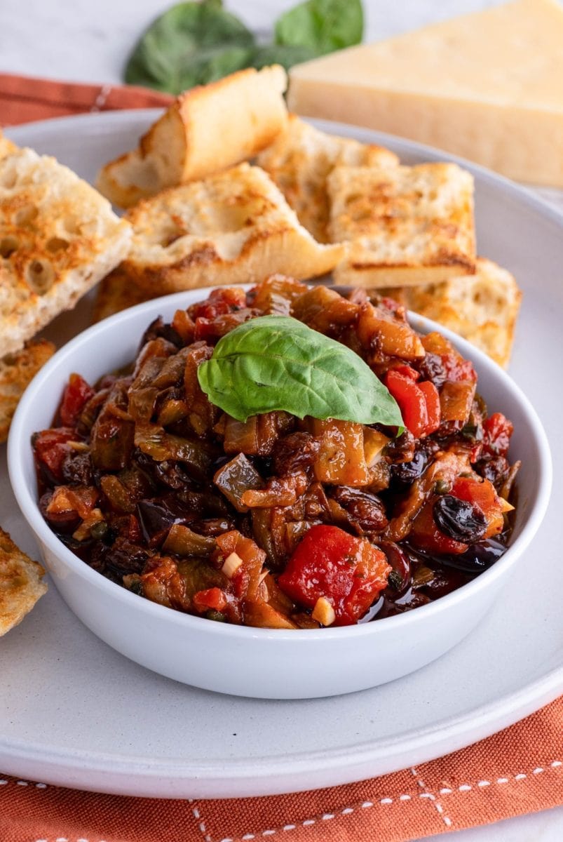 Bowl of eggplant caponata on a plate with toasted baguette, ready to serve.