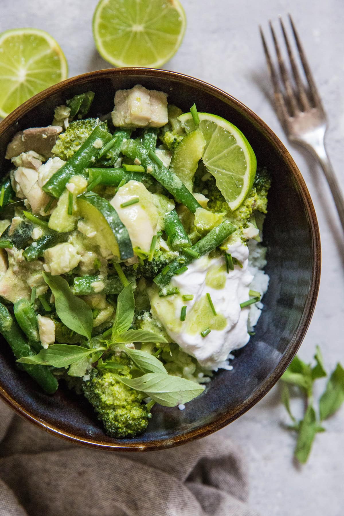 top down photo of green fish curry in a blue bowl with sliced of lime and Thai basil with a fork and napkin to the side